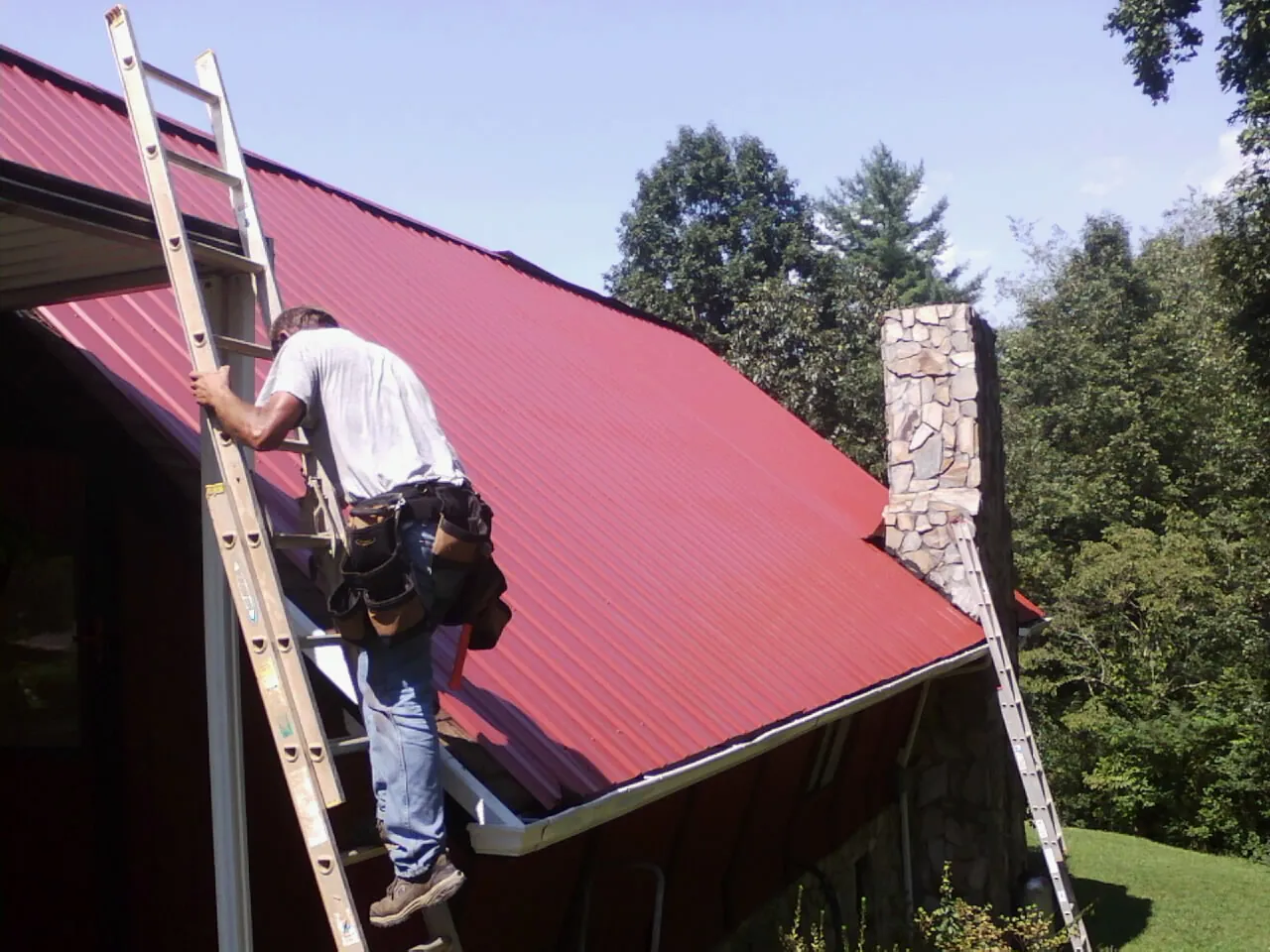 Roofer on a ladder beside a red metal roof and stone chimney — Soffit & Fascia Repair by Roofing Brothers in Middle Haddam